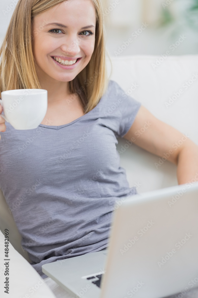 Fototapeta premium Smiling woman with coffee cup using laptop on sofa