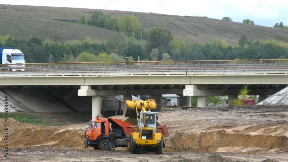 Dredger puts earth in dump truck in front of automobile bridge Dredger puts earth in dump truck in front of automobile bridge