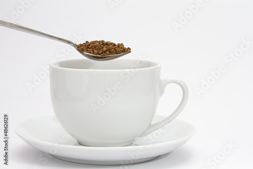 Spoon of instant coffee granules above white cup and saucer