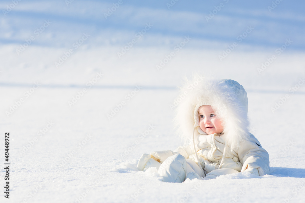 Baby in white suit sitting in snow field on sunny winter day Baby in white suit sitting in snow field on sunny winter day