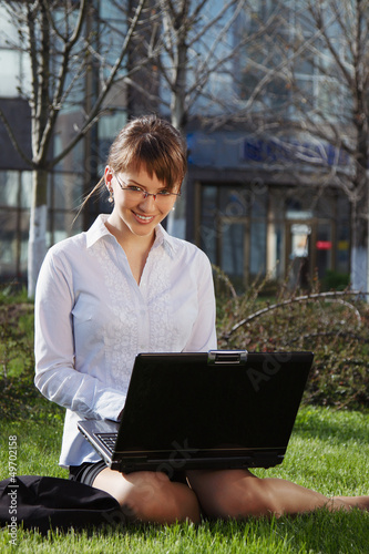 Woman lying on grass with laptop with business building on back
