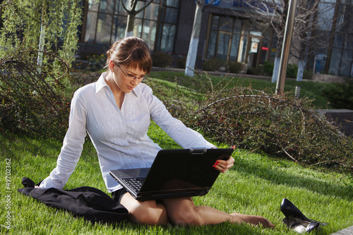Woman lying on grass with laptop with business building on back