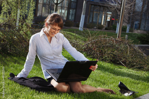 Woman lying on grass with laptop with business building on back