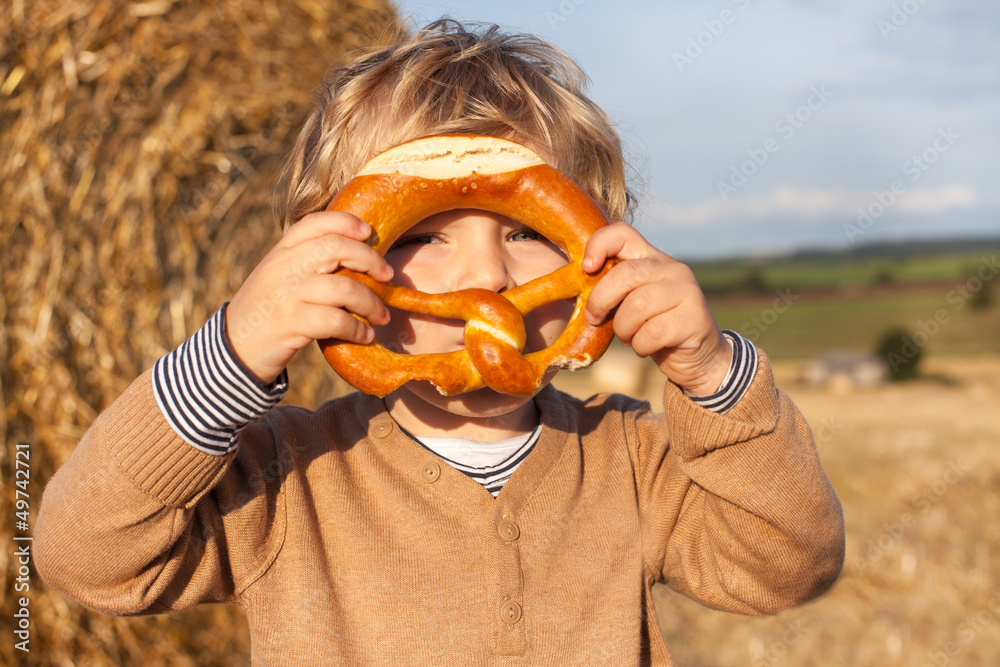 Fototapeta premium Cute toddler eating German pretzel on goden hay field