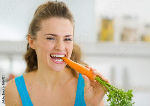 Happy young woman eating carrot in kitchen