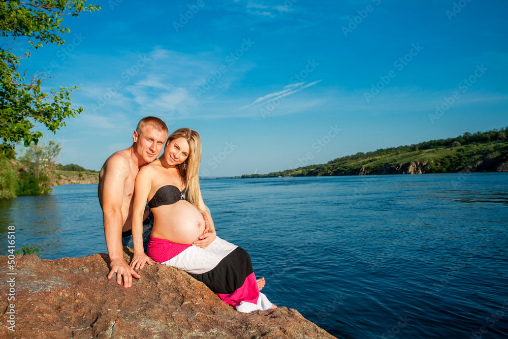 Pregnant woman and her husband embraces near water