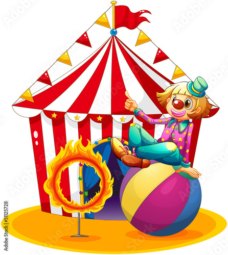 A clown sitting  above a ball beside a ring of fire in front of