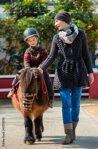 Mother riding her son on a pony wearing protective helmet
