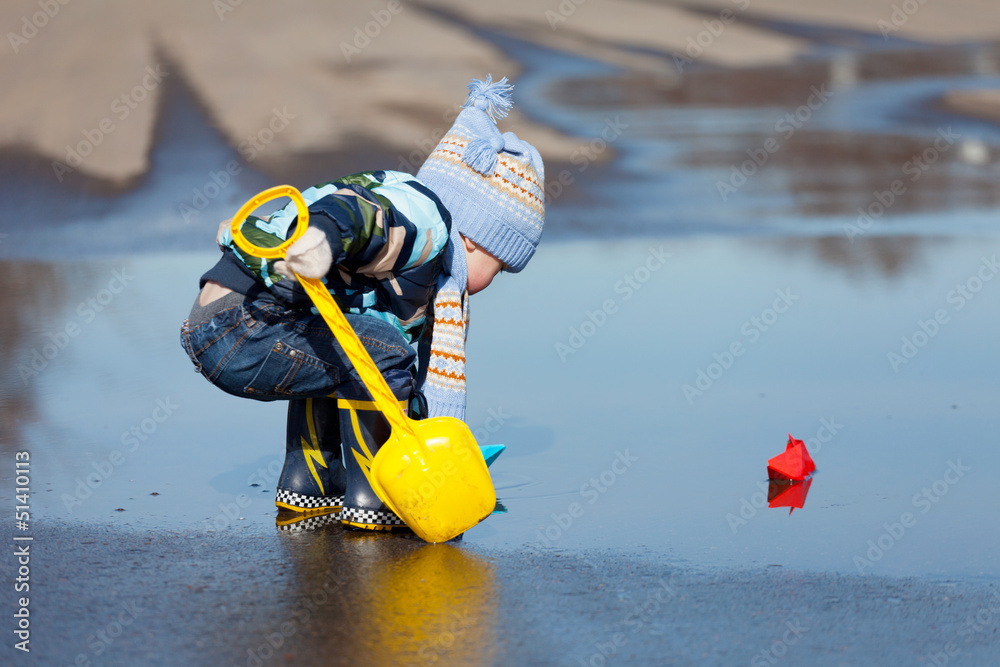 Little boy plays with paper ships in a spring puddle
