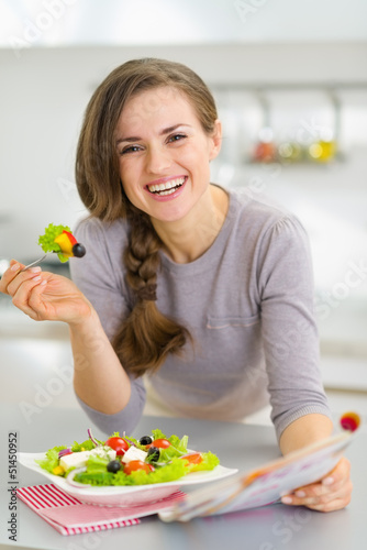 Smiling young housewife eating fresh salad and reading magazine