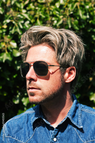 Man with blonde hair in front of green foliage. Wearing sunglass