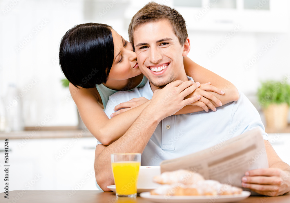 Girl kisses eating boyfriend who sits at the kitchen table 