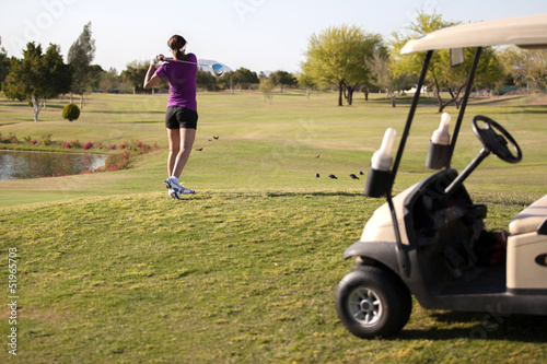Young female golfer teeing off next to a golf cart