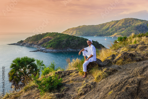 The man in a white suit and hat sitting on a rock on the sea ba