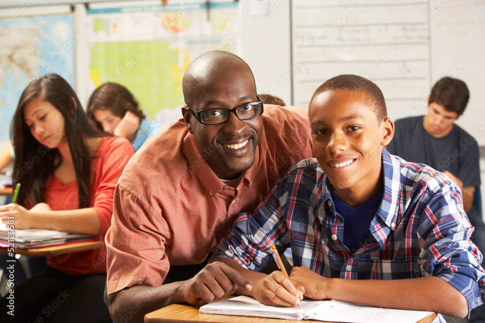 Fototapeta premium Teacher Helping Male Pupil Studying At Desk In Classroom