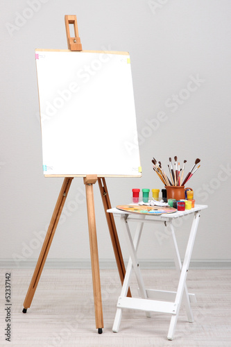 Wooden easel with clean paper and art supplies in room