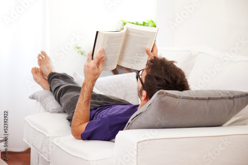 Young man stretching comfortably on couch and reading a book