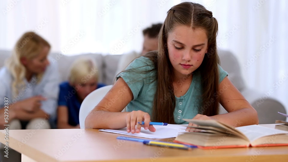 Daughter doing her homework with family on couch behind Daughter doing her homework with family on couch behind