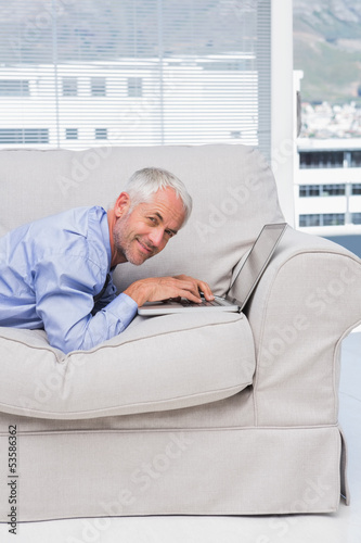 Businessman lying on sofa with laptop smiling at camera