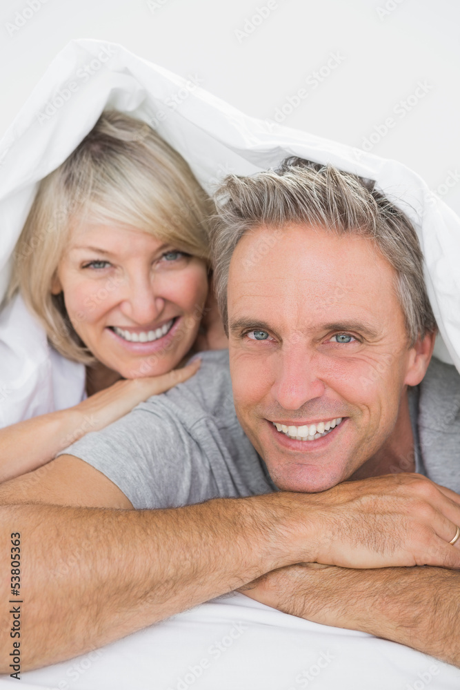 Couple smiling under the covers at the camera Couple smiling under the covers at the camera