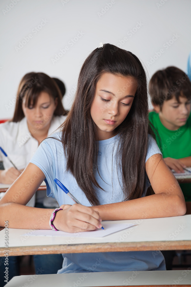 Female Student Writing Paper At Desk In Classroom Female Student Writing Paper At Desk In Classroom