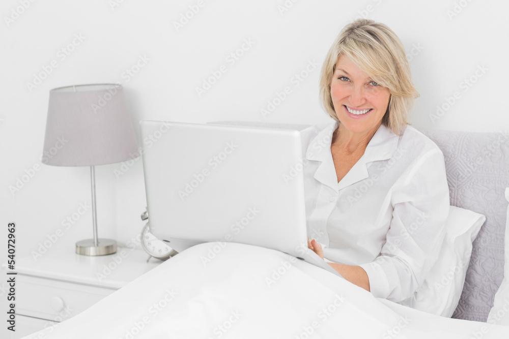 Happy woman sitting in bed with laptop looking at camera Happy woman sitting in bed with laptop looking at camera