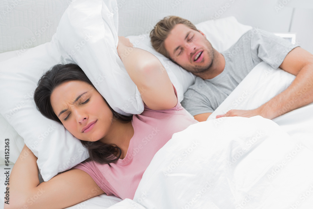 Woman covering her ears with pillow while her husband is snoring Woman covering her ears with pillow while her husband is snoring