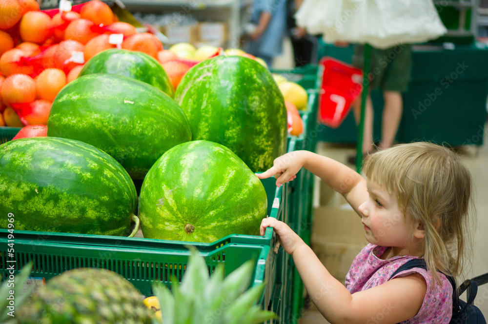 Fototapeta premium Adorable girl with backpack select watermelon in supermarket