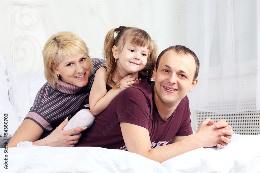 Little girl, father and mother lie on white double bed and smile Little girl, father and mother lie on white double bed and smile