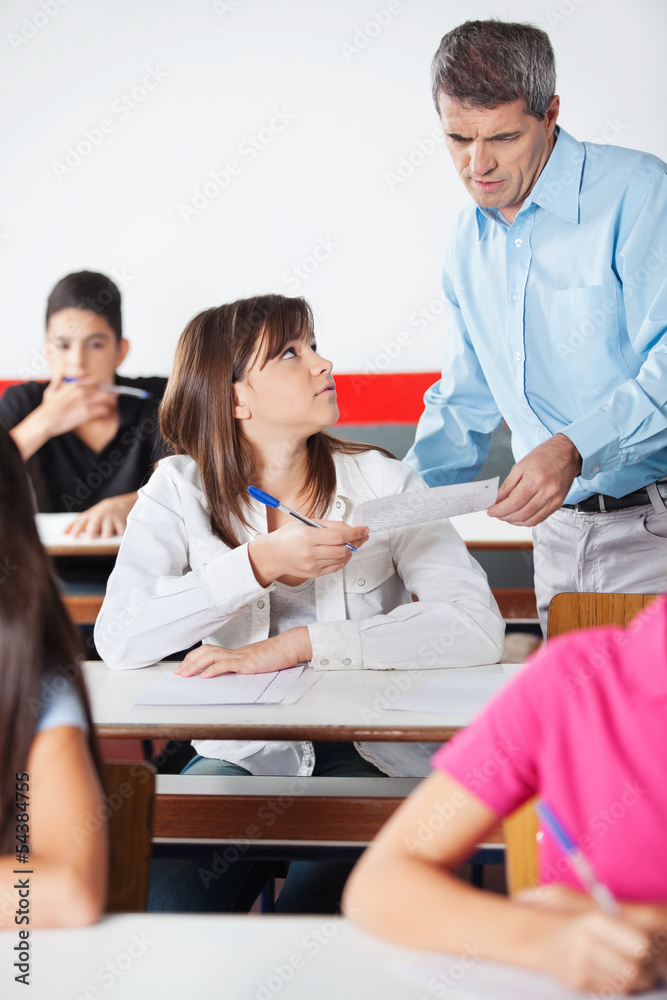 Female Student Showing Paper To Teacher At Desk Female Student Showing Paper To Teacher At Desk