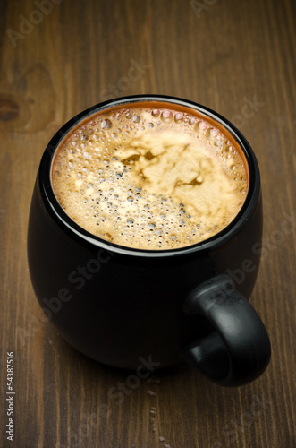 mug of black coffee with foam on a wooden table, top view