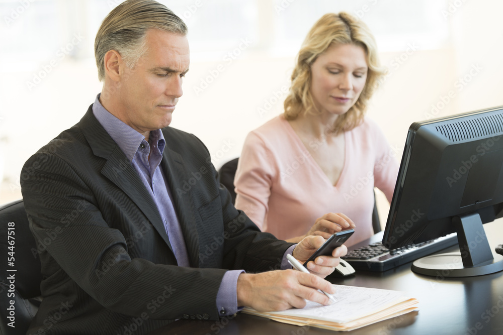 Businessman Using Mobile Phone While Colleague Working At Desk Businessman Using Mobile Phone While Colleague Working At Desk