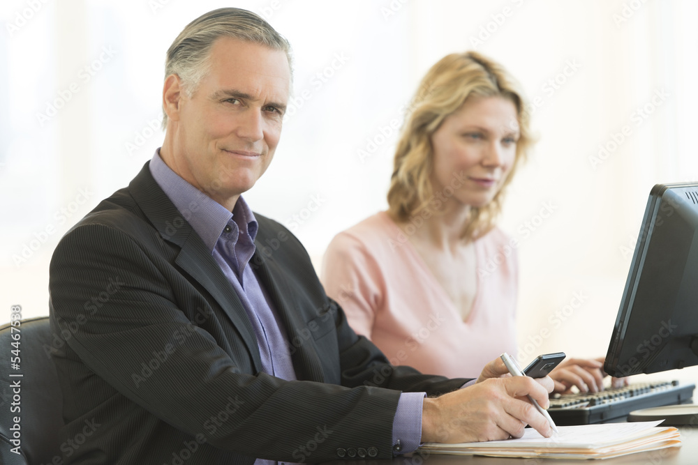 Confident Businessman Holding Pen And Mobile Phone In Office Confident Businessman Holding Pen And Mobile Phone In Office