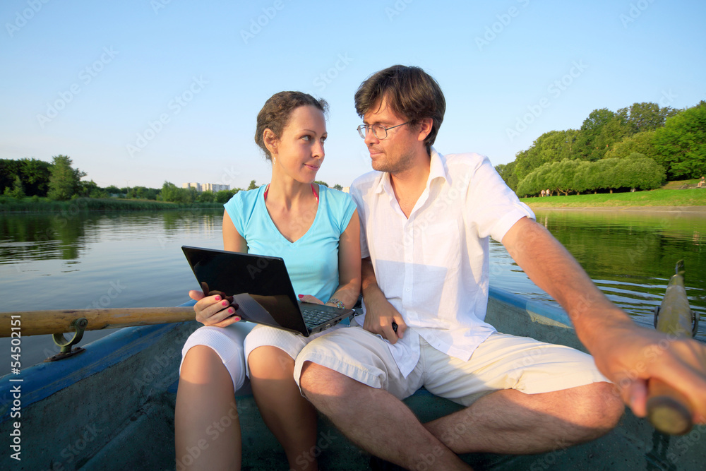 Young couple floating down river on boat with oars with laptop Young couple floating down river on boat with oars with laptop