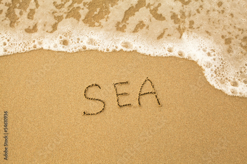 Sea - written by hand in sand on a sea beach, with a soft wave.