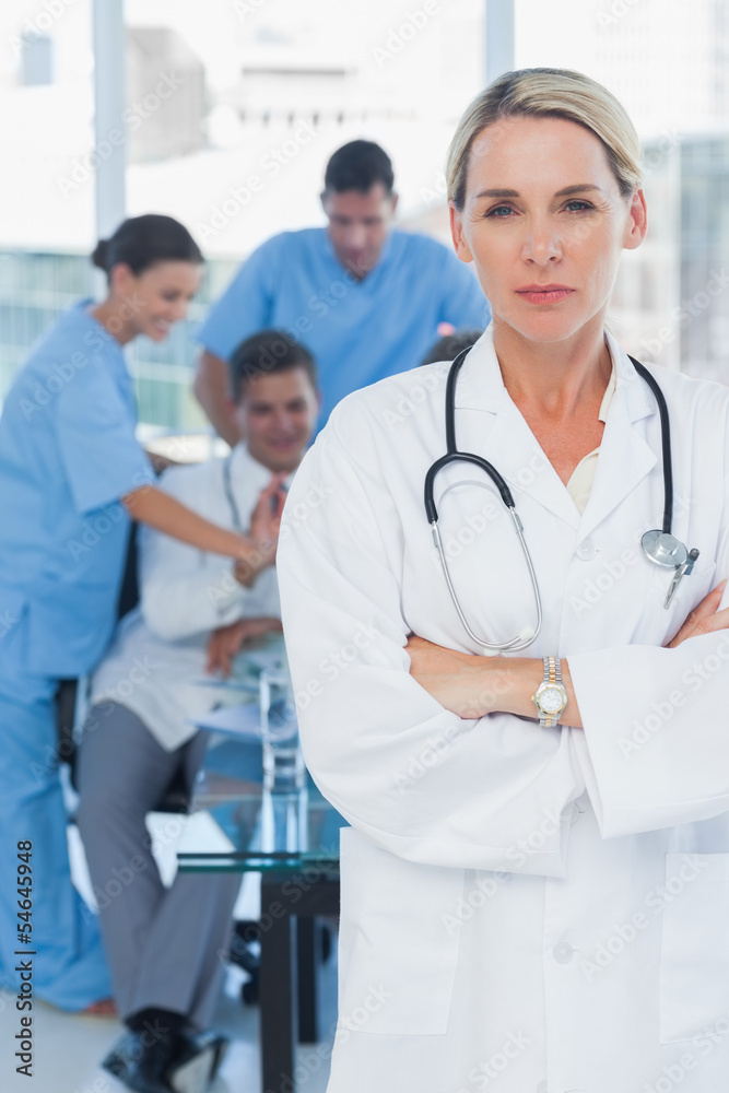 Serious blond doctor posing with colleagues in background Serious blond doctor posing with colleagues in background