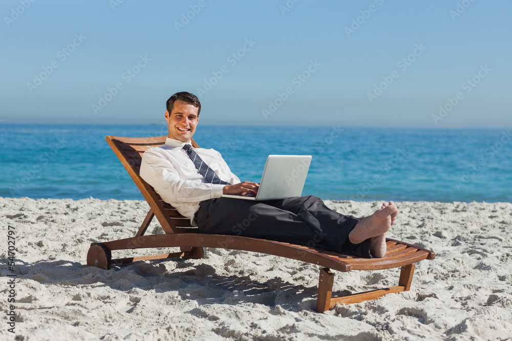 Smiling young businessman lying on a deck chair with his laptop