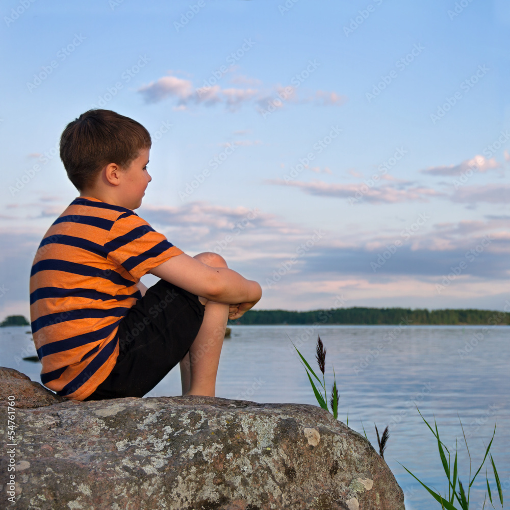 Fototapeta premium Boy sitting on a large rock watching a seaside landscape