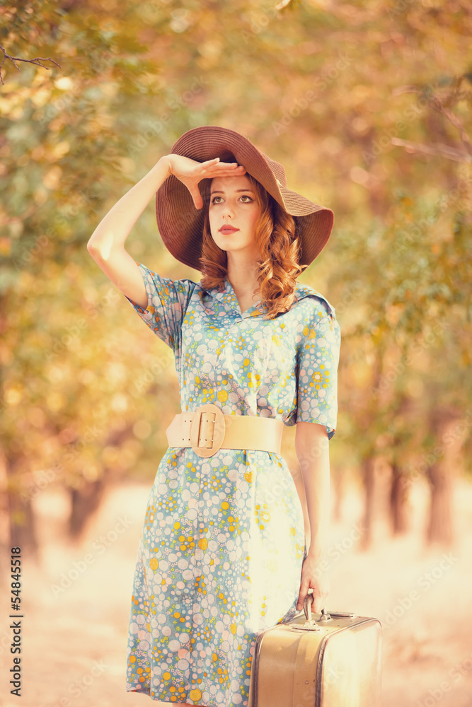 Obraz premium Redhead girl with suitcase at tree's alley.