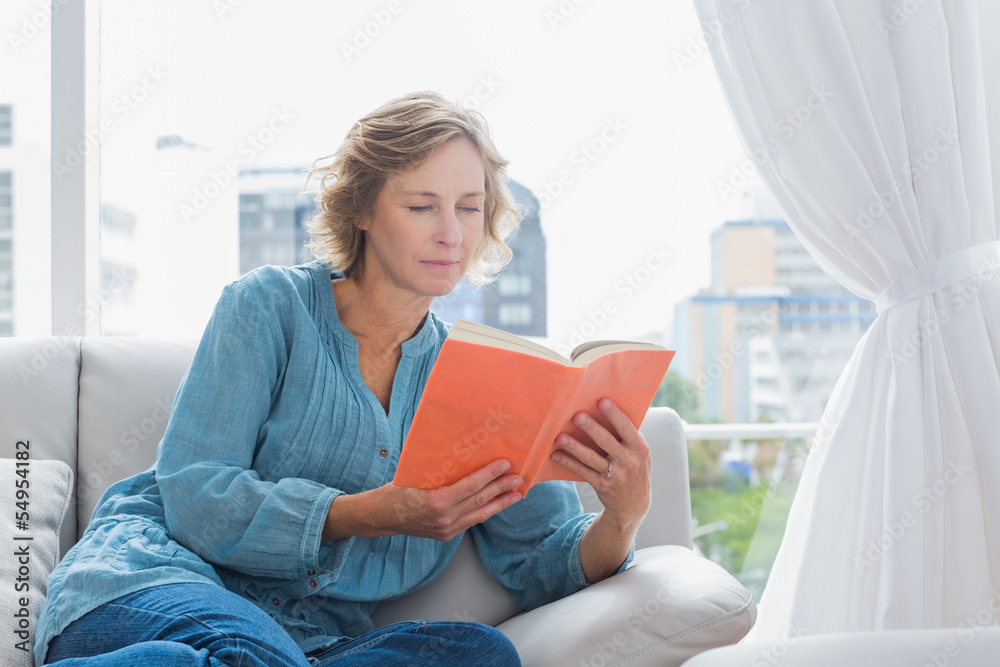 Cheerful blonde woman sitting on her couch reading a book Cheerful blonde woman sitting on her couch reading a book