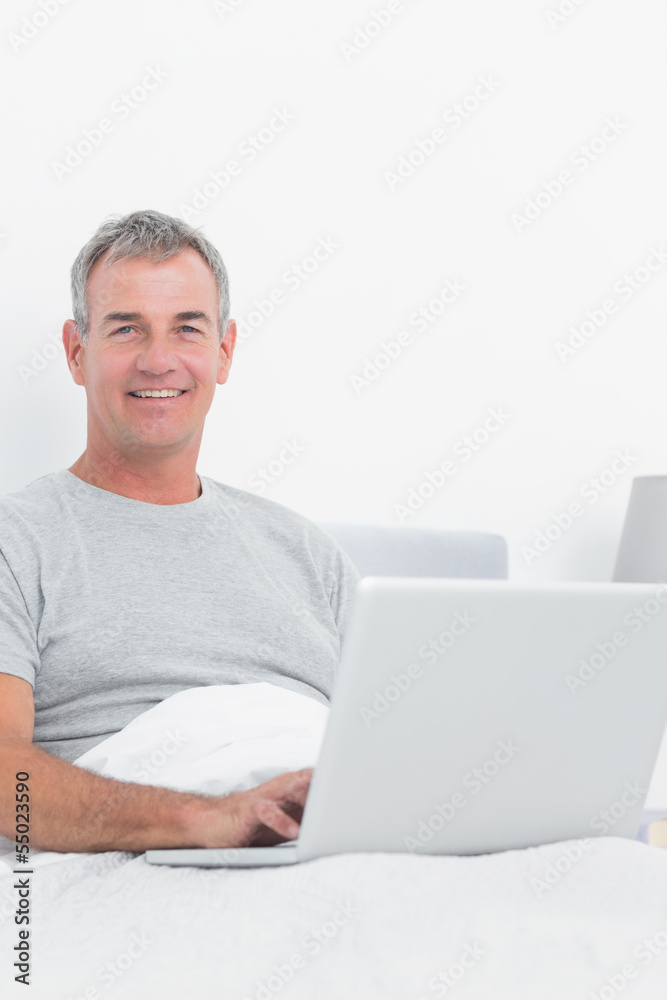 Cheerful grey haired man using his laptop in bed Cheerful grey haired man using his laptop in bed