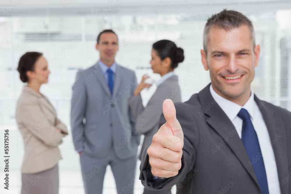 Cheerful manager showing thumb up with employees in background Cheerful manager showing thumb up with employees in background
