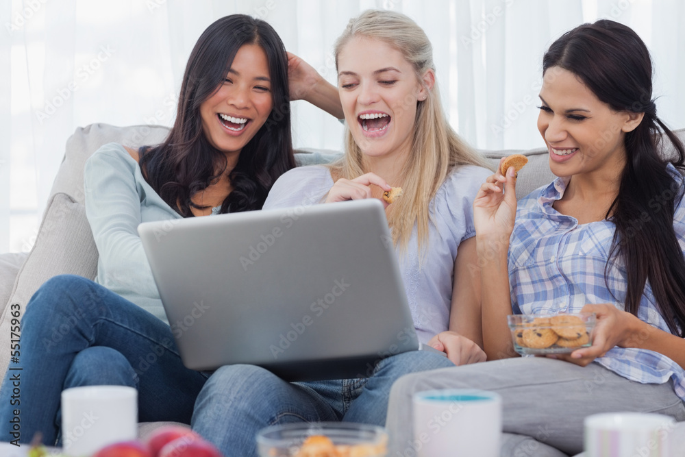 Laughing friends looking at laptop together and eating cookies Laughing friends looking at laptop together and eating cookies