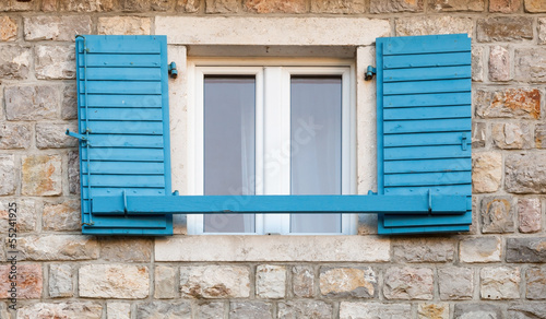Wooden window with blue open jalousies in old gray stone wall