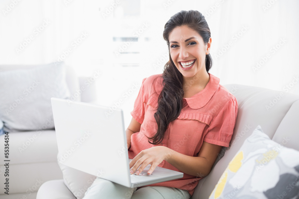 Naklejka premium Smiling brunette sitting on her sofa using laptop