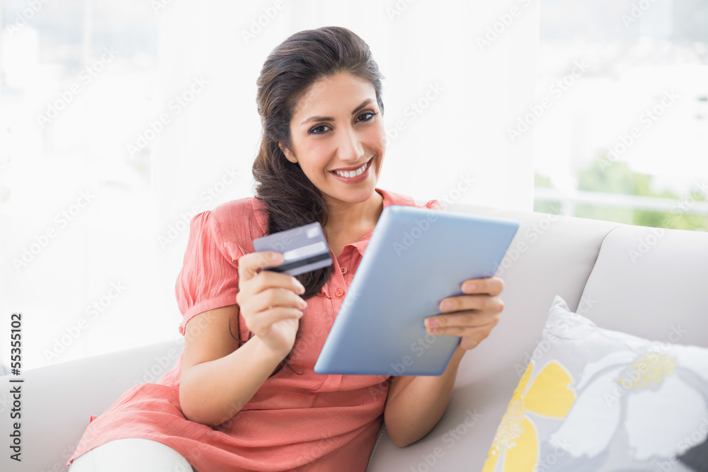 Smiling brunette sitting on her sofa using tablet to shop online Smiling brunette sitting on her sofa using tablet to shop online