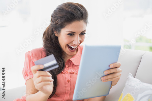 Ecstatic brunette sitting on her sofa using tablet to shop onlin