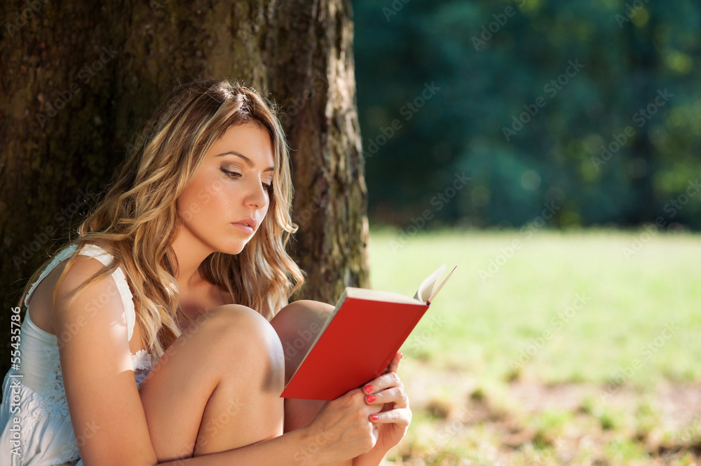 Beautiful young woman reading a book under a tree in the park.