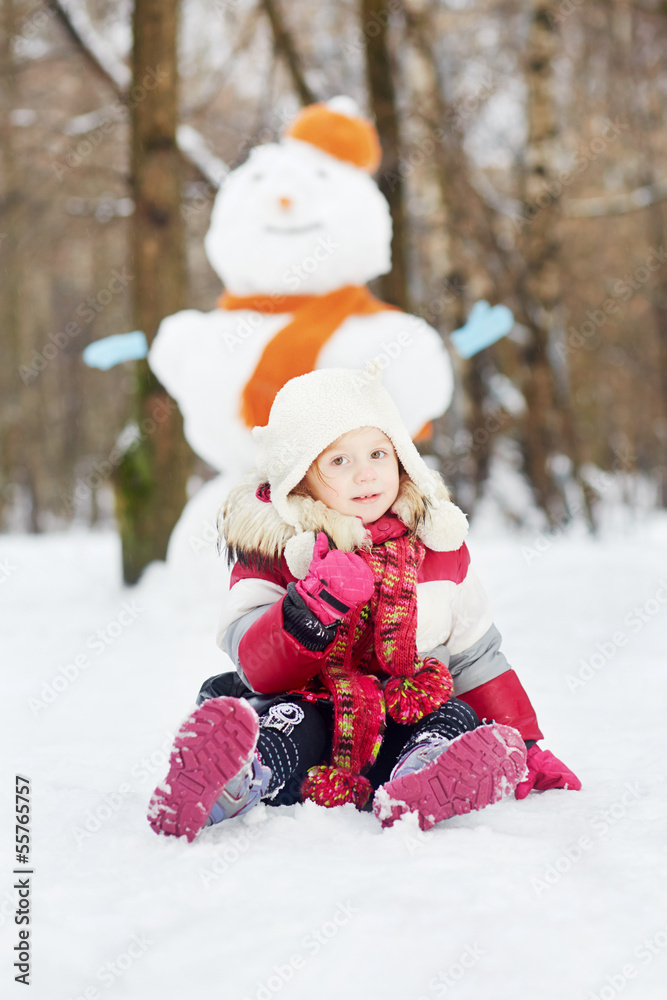 Little girl sits on snow in front of big snowman in winter park,