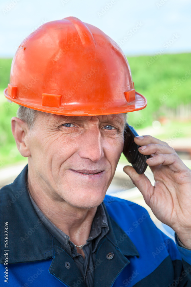 View of mature manual worker in orange hardhat calling on phone View of mature manual worker in orange hardhat calling on phone